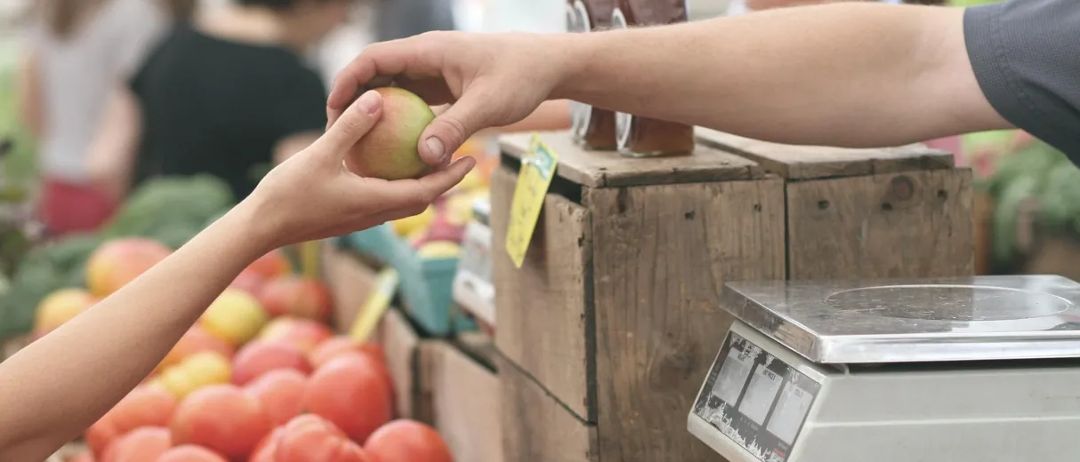 Zwei Hände tauschen einen grünen Apfel über einer bunten Auslage frischer Marktprodukte.